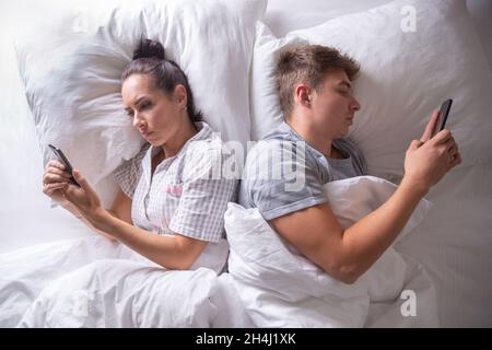 Couple separated in bed by digital technology as they both lie backs to each other looking at their cell phones. Stock Photo