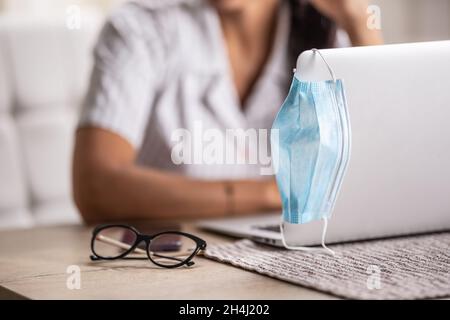 Detail of a face mask hung on a laptop and glasses put aside as woman keeps on working remotely after pandemic. Stock Photo