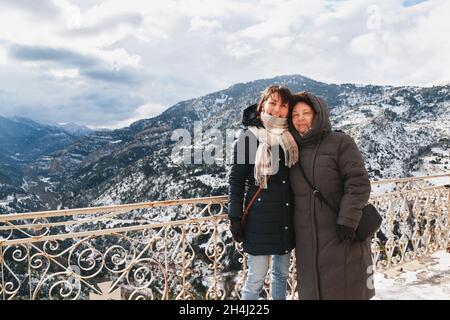 Winter portrait of hugging and smiling mother and daughter with splendid snow-covered mountains view in background, from Mega Spileon Monastery terrace at Vouraikos gorge. Winter vacation. Greece Stock Photo
