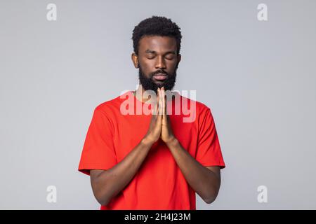African American man in white shirt, closing eyes, holding hands in pray and making wish, praying or hoping for better over gray background Stock Photo