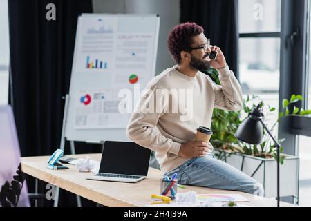 Positive african american businessman in eyeglasses talking on smartphone and holding takeaway drink near laptop in office Stock Photo
