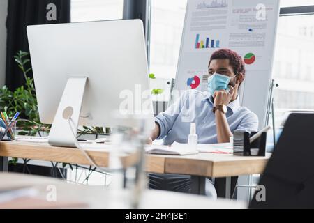 African american businessman in medical mask using computer near papers and hand sanitizer in office Stock Photo