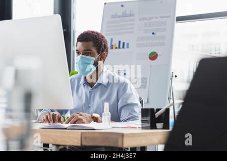 African american businessman in protective mask using computer near blurred flip chart in office Stock Photo