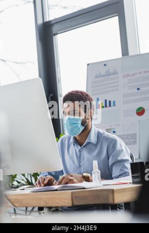 African american businessman in medical mask using laptop near hand sanitizer in office Stock Photo