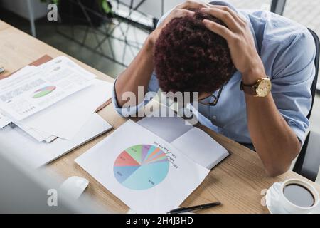 High angle view of upset african american businessman touching head near papers and coffee on office table Stock Photo