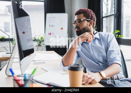 African american businessman in eyeglasses looking at computer near coffee to go and notebook in office Stock Photo
