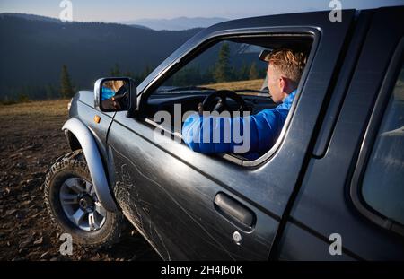 Close-up of driver in cabin of black SUV driving on stone roads of mountain hills. Extreme car adventure in the mountains. On the background mountain beskids covered by green forests. Stock Photo