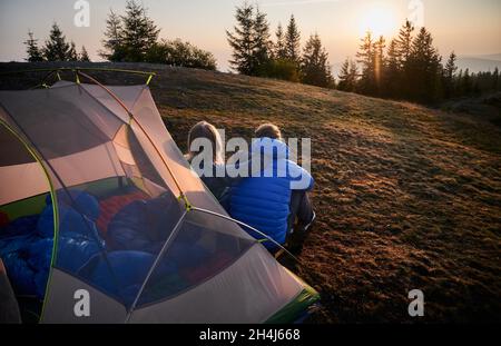 Back view of two travelers man and woman sitting on grass in front of tent set up on mountain hill, early in the morning and contemplating on sunny sky. Concept of hiking, camping and relationships. Stock Photo