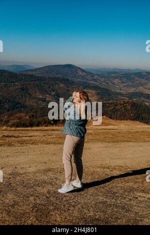A young woman in a warm suit and a denim jacket rejoices in the mountains. Autumn in the mountains. Happy girl. Nature and travel. Vertical photo. Stock Photo
