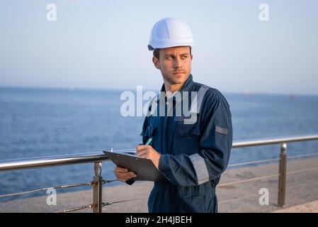 Serious young male engineer in hardhat and protective clothes looking away while inspecting sea port and taking notes on clipboard Stock Photo