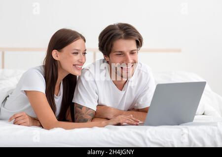 Cheerful young caucasian couple lie on bed with laptop have video call, watch video, chatting, searching Stock Photo