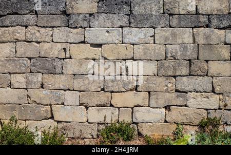 Texture, background, pattern. Ancient dry built stone brick wall. Irregular stones broken and ruined by time. Weeds grow low. Rough and dirty surface. Stock Photo