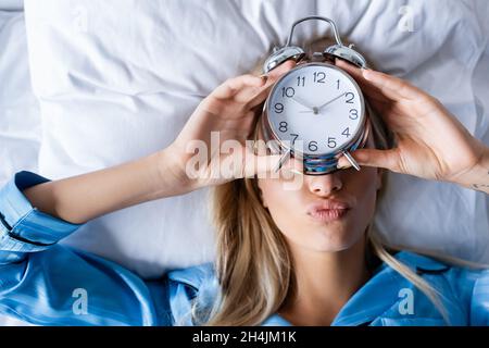 top view of blonde woman holding retro alarm clock and pouting lips while lying on bed Stock Photo