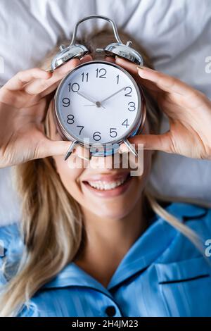 top view of cheerful woman holding retro alarm clock and covering face while lying on bed Stock Photo