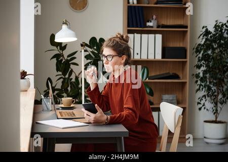 Young woman sitting at the table with laptop and using mobile phone at her work at office Stock Photo
