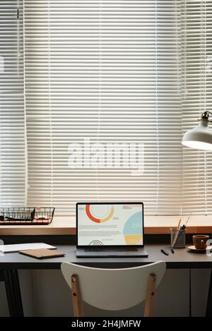 Image of laptop with financial charts on the screen on the table in front of the big window at office Stock Photo