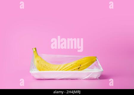 Fruits in plastic packaging from the supermarket are minimal. Banana in cellophane and non-degradable plastic on a blue background. Biodegradable product packaging, environmental protection, nature pollution. High quality photo Stock Photo