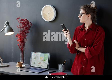 Young businesswoman in eyeglasses looking away and thinking about something while using her mobile phone at modern office Stock Photo