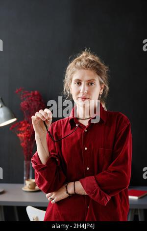 Portrait of young businesswoman in red shirt taking off her eyeglasses and looking at camera while standing at office Stock Photo