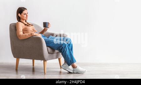 Full length of millennial woman drinking fresh coffee, having break in comfy armchair against white studio wall, panorama with empty space. Calm young Stock Photo