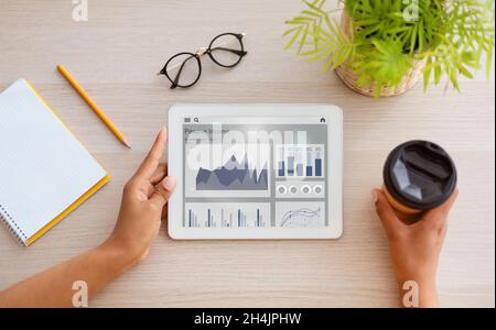 Hands of young african american woman hold cup of coffee and tablet with digital charts on tablet screen at workplace, collage, top view. Modern succe Stock Photo