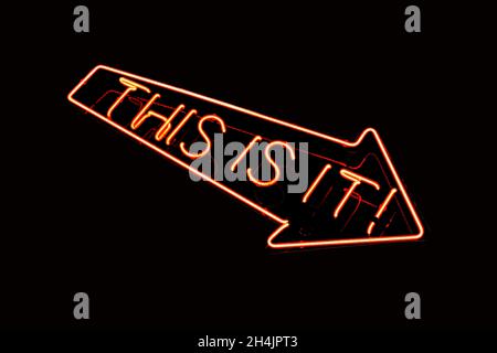 Neon sign in red with text saying THIS IS IT. Downward pointing arrow isolated on black background. Stock Photo