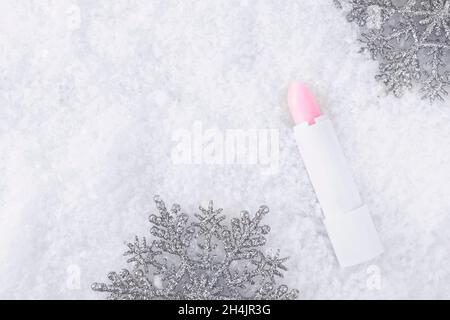 A white tube of pink lipstick balm to protect and nourish lips. Winter season cosmetics. Mock up container near with snowflakes, in the snow. Christma Stock Photo