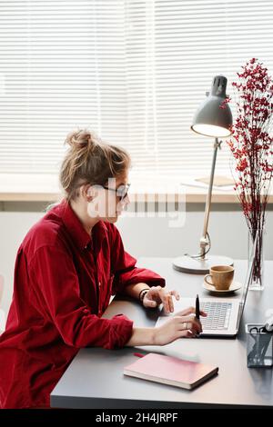 Young woman in eyeglasses sitting at the table and studying online using her laptop at home Stock Photo