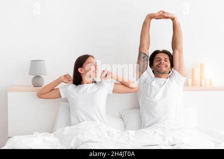 Happy caucasian millennial wife and husband woke up and stretching body sitting on bed in white bedroom interior, copy space. Good morning together, c Stock Photo