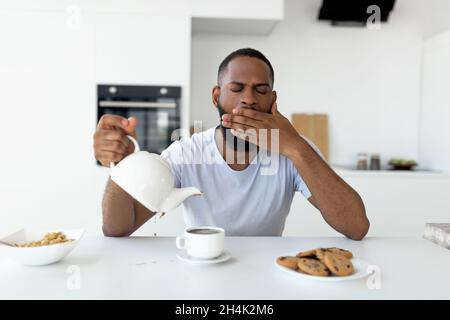 Lack Of Sleep Concept. Portrait of tired young African American man yawning while sitting at dining table in kitchen, holding teapot pouring coffee aw Stock Photo
