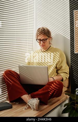 Young woman in eyeglasses sitting on windowsill and communicating online using her laptop Stock Photo