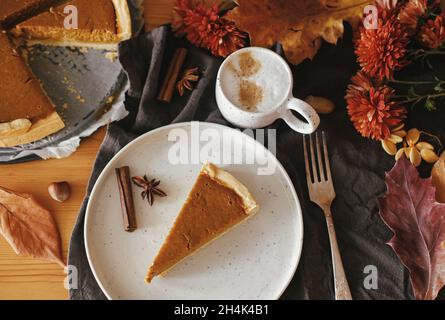 Happy Thanksgiving. Pumpkin pie slice on modern plate and hot coffee on rustic table with linen napkin, autumn flowers and leaves, anise and cinnamon. Stock Photo