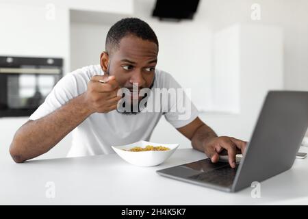 Gadget Addiction. Portrait of black guy having breakfast in kitchen at home, eating healthy cereal, using and looking at laptop screen, working remote Stock Photo