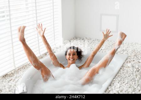 Silly young woman having fun in bubble bath, raising her arms and legs up, being silly and playful at home. Millennial female enjoying domestic beauty Stock Photo