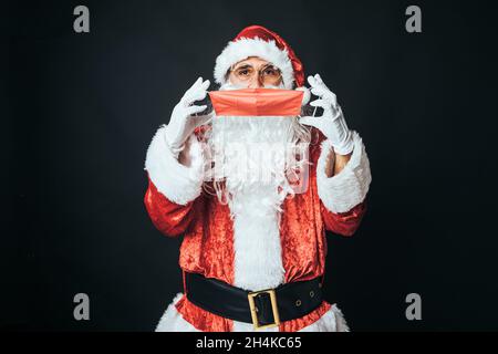 Man dressed as Santa Claus putting on a red mask to avoid covid infection, on a black background. Christmas concept, Santa Claus, gifts, celebration.