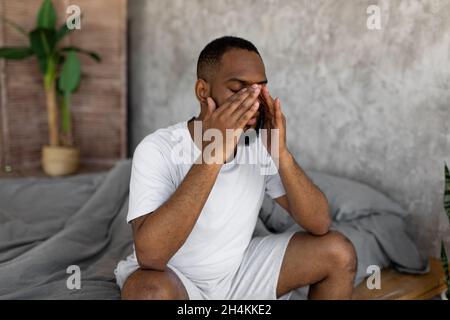 Young black man suffering from headache or migraine, rubbing eyes after waking up. Stressed guy sitting on bed with painful face expression feeling te Stock Photo