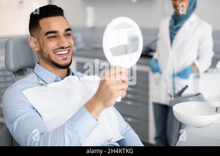 Happy Handsome Arab Man Looking To Mirror After Teeth Treatment In Modern Clinic, Middle Eastern Male Patient Sitting In Chair In Stomatological Cabin Stock Photo