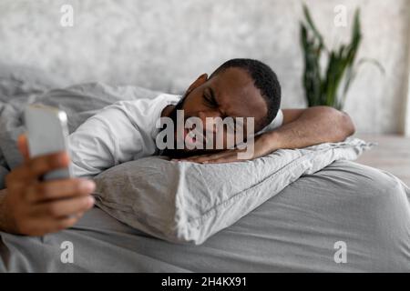 Bad News And Oversleep. Shocked confused black man wakes up with anxiety, surprised upset angry young guy turned off alarm clock on smartphone lying o Stock Photo