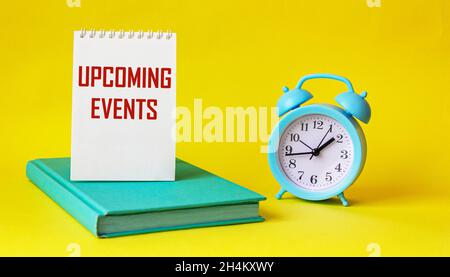 Conceptual message UPCOMING EVENTS in notepad and on yellow background, next to alarm clock and green diary. The best view of the modern workplace. Stock Photo