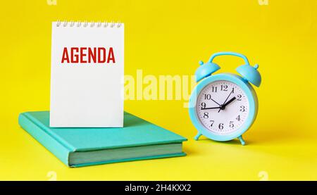 The word AGENDA is written on a notebook that lies on a green diary, next to an alarm clock on a yellow background Stock Photo