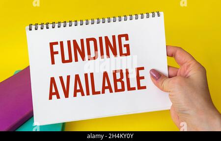 Funding Available written on a notepad in a woman's hand on a yellow background, next to colored notepads. Sponsor financing business start-up project Stock Photo