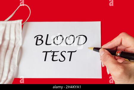 Medical concept. The phrase BLOOD TEST is written by a female hand on a notebook, next to a medical mask on a red background Stock Photo