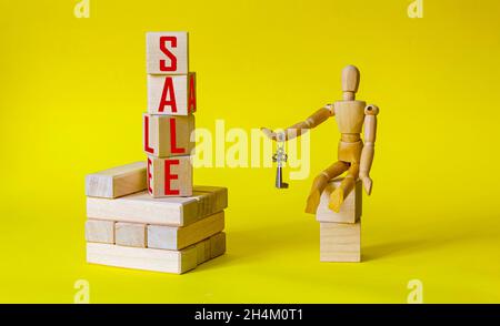 The word sale is written on wooden blocks. The wooden model of a man sits on the cubes and holds the keys. Beautiful yellow background. Business and s Stock Photo