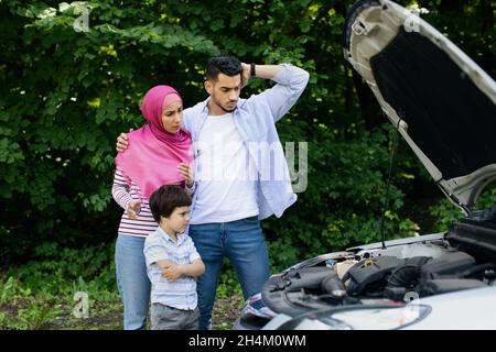 Confused Muslim Family With Little Son Standing Near Broken Car Outdoors, Upset Middle Eastern Parents And Male Child Suffering Troubles With Their Ve Stock Photo