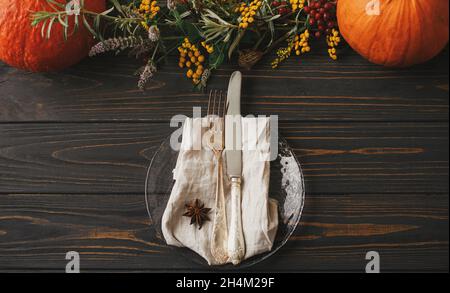 Stylish Thanksgiving dinner table setting. Modern plate with vintage cutlery, linen napkin with anise on wooden table with pumpkins and autumn flowers Stock Photo