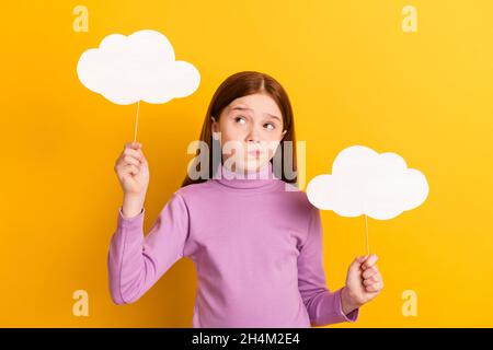 Photo of uncertain unsure doubtful thoughtful little girl hold paper clouds look copyspace isolated on yellow color background Stock Photo