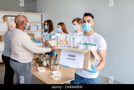 Young male volunteer holding cardboard box for donation and posing to camera while group of colleagues giving elderly people humanitarian aid, working Stock Photo