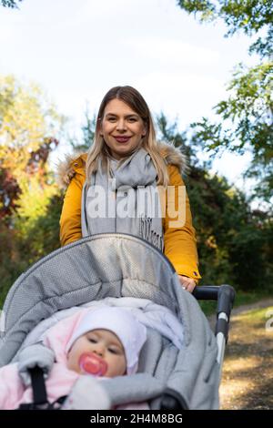 Picture of young cheerful caucasian lady walking her baby with a stroller outdoor. Baby having a pacifier Stock Photo