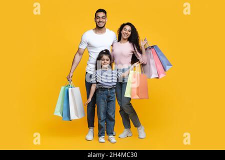 Seasonal Sales. Portrait Of Happy Arab Family With Little Daughter Holding Lots Of Bright Shopping Bags, Middle Eastern Parents And Child Posing Over Stock Photo