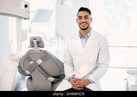 Portrait Of Smiling Middle Eastern Male Dentist Doctor Sitting At Workplace In Modern Clinic, Handsome Arab Stomatologist Man Wearing Uniform Posing A Stock Photo